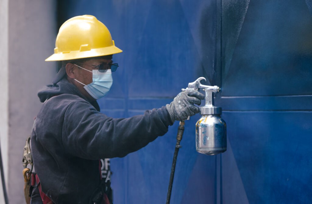 Operario pintando con pistola en cabina; ventilación industrial y extracción localizada para control de vapores y neblina de pintura