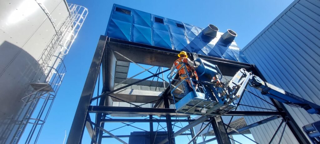 Técnico realizando instalación y mantenimiento en altura en colector de polvos tipo baghouse; uso de plataforma elevadora y EPP para filtración industrial