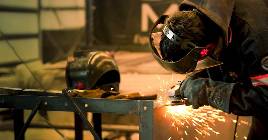 Operario esmerilando metal con chispas; extracción localizada y ventilación industrial para controlar humos metálicos, polvo y partículas en taller